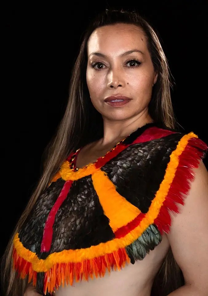 A studio portrait of Dr. Michelle Manu shows her looking directly at the camera with a calm expression. She has long, straight, light brown hair flowing over her shoulders. She is wearing a striking traditional-looking cape or collar made of black, red, and orange feathers, with some green feathers at the bottom. Her shoulders and chest are bare, and the background is solid black.