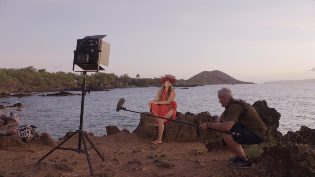 A film set on a rocky coastline at dusk. Dr. Michelle Manu is seated in the center, wearing a red haku lei (head lei) and a red dress, looking towards a camera (not visible). To her left, a large square light panel on a tripod illuminates her. To her right, a man with gray hair, wearing a green shirt and dark shorts, crouches while holding a boom microphone towards her. The background features the ocean with gentle waves, a distant island or hill, and lush green foliage along the shore. The sky is a pale orange and pink, indicating sunset.