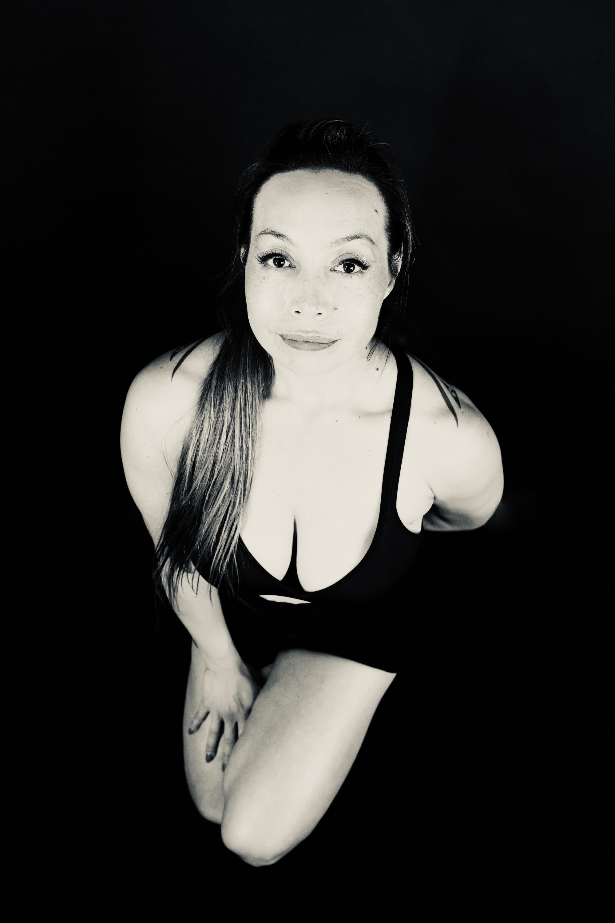 A high-angle, black and white portrait of Dr. Michelle Manu shows her on her knees, looking up directly at the viewer with a slight smile. She has light skin and long, light-colored hair flowing over her shoulders. She is wearing a dark, possibly black, bra top and shorts. The background is completely dark.