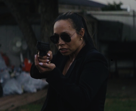 A dramatic mid-shot of Dr. Michelle Manu shows her aiming a handgun with both hands, looking intently forward. She has dark hair pulled back into a ponytail, is wearing dark sunglasses, and a black blazer over a dark top. Her mouth is slightly open, suggesting concentration or exertion. The background is blurred, showing what appears to be an outdoor setting with a tree and some indistinct objects.