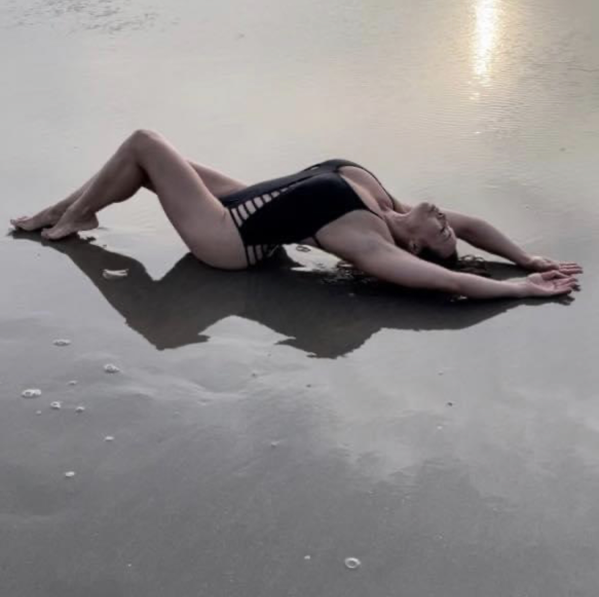 A full-body shot of Dr. Michelle Manu, a woman with dark hair, lying on her back on wet sand at what appears to be sunset or sunrise. She is wearing a dark, possibly black, one-piece swimsuit with cutout details on the sides. Her knees are bent with her feet flat on the ground, and her arms are stretched out overhead on the sand. Her head is tilted back, looking upwards. The wet sand reflects the light, and a faint glow from the sun is visible on the water in the background.