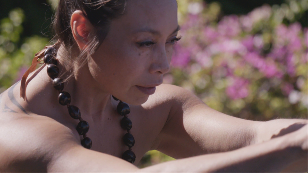 A close-up outdoor shot of Dr. Michelle Manu, a woman with light skin and dark hair pulled back from her face. She is looking down and to her right with a serious expression. She wears a dark, possibly kukui nut, lei around her neck. Her bare shoulder with a faint tattoo and arm are visible in the foreground. The background is softly blurred with green foliage and bright pink flowers.