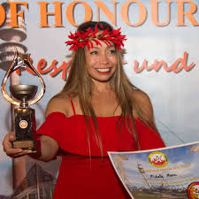 A medium shot of Dr. Michelle Manu, a woman with long, straight brown hair, smiling brightly as she holds up a golden trophy in her left hand and a certificate in her right. She is wearing a vibrant red off-the-shoulder dress and a red floral headpiece. Behind her, a banner with the words "OF HONOUR" and "und" in large orange letters is visible, along with a blurred image of a structure that resembles a lighthouse.