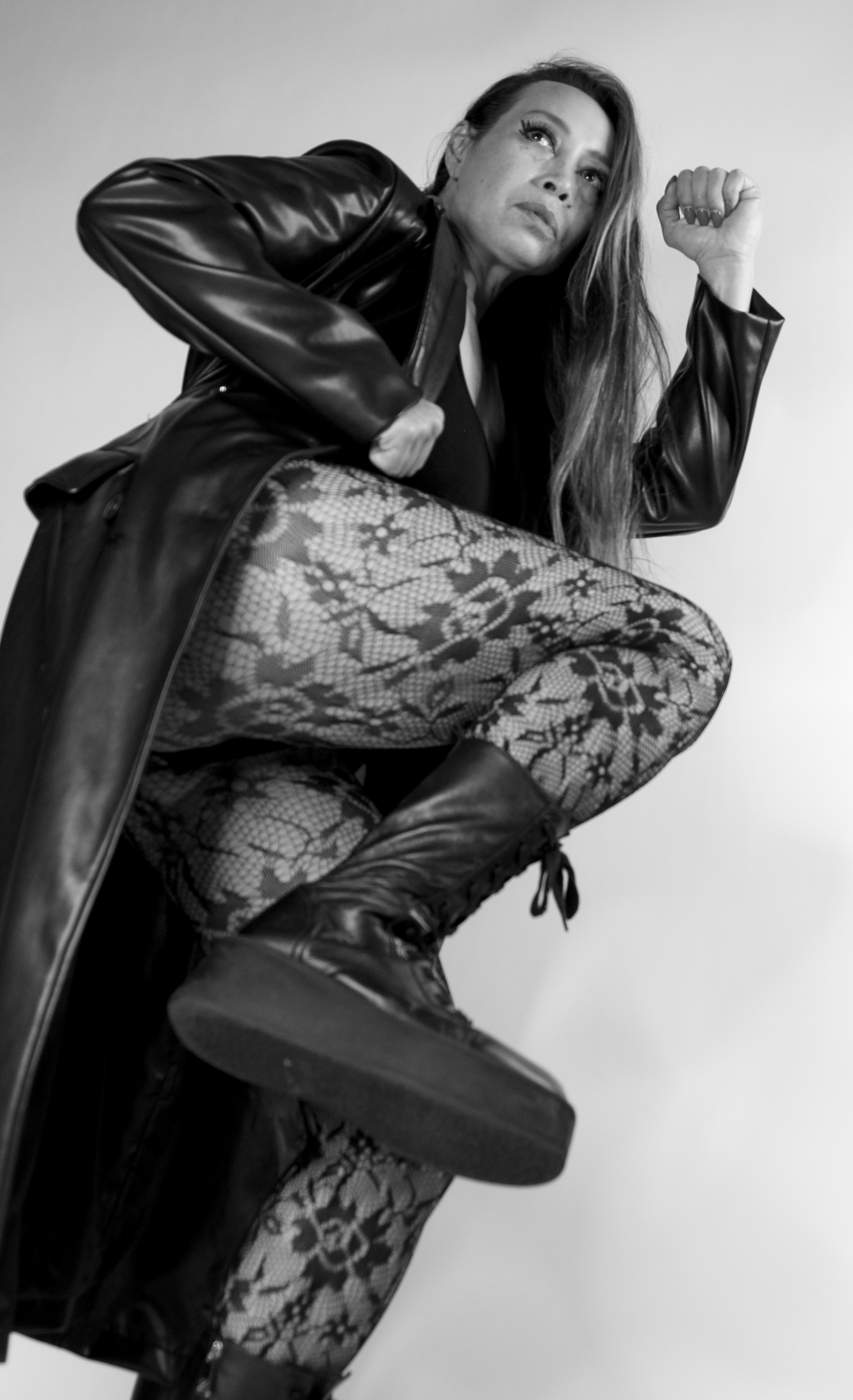 Dr. Michelle Manu in a dynamic pose, wearing a long leather coat, patterned tights, and combat boots. She is lifting one leg high as if stepping forward or striking, looking intensely at the camera with one arm bent upward. The image conveys strength and confidence.