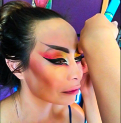 Michelle Manu sits still with her eyes closed as another person applies bold, theatrical makeup to her face. The makeup features sharp, exaggerated lines in vibrant red, yellow, and black, forming a dramatic and stylized design. Her hair is tied in a bun, and the setting includes a purple wall and other colorful background elements, suggesting a preparation area for a performance or cultural event.