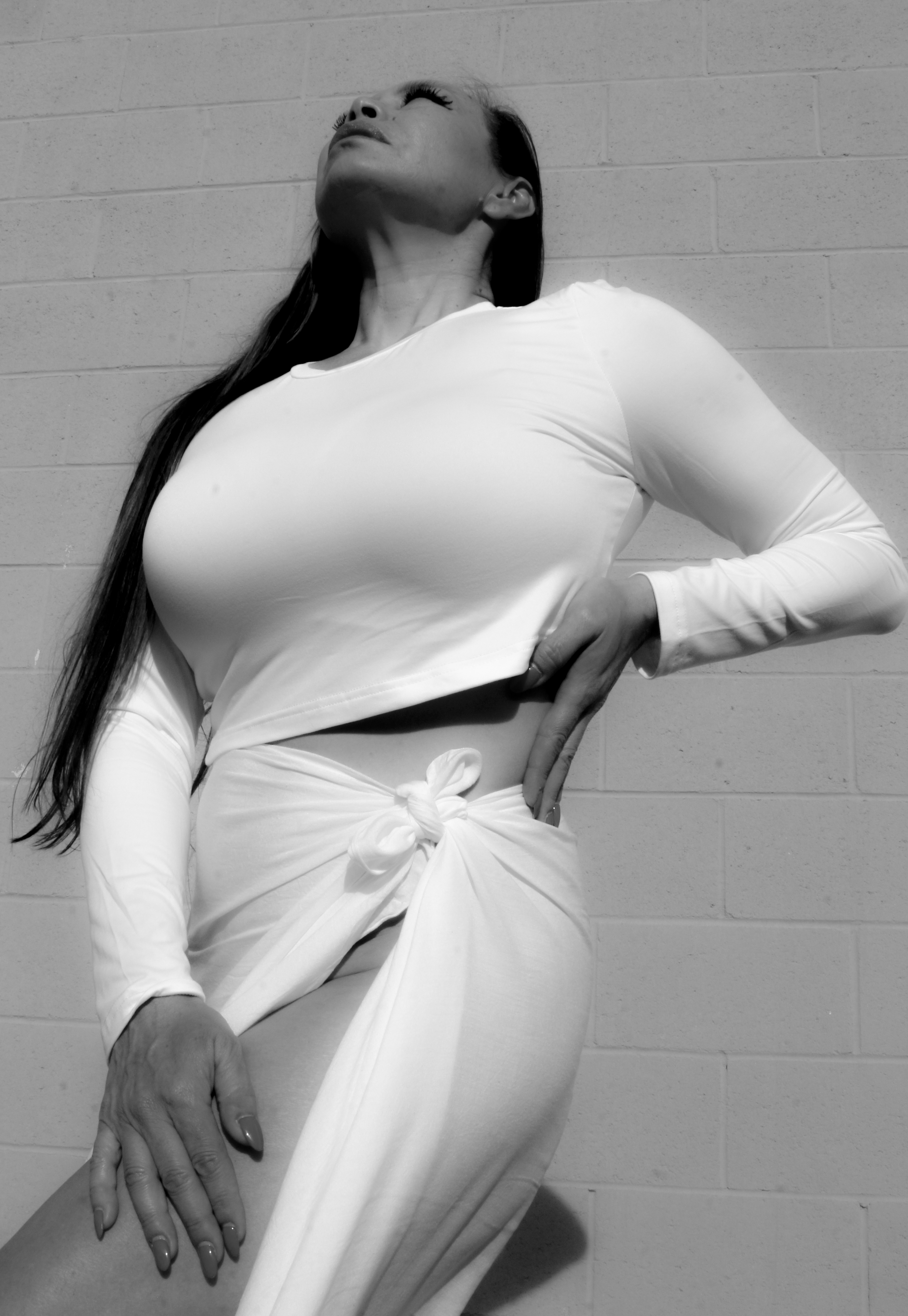 A black and white, low-angle shot features Dr. Michelle Manu, a woman with long, dark hair, posing dramatically. She is looking upwards with her chin raised, illuminated by strong light from above. She wears a long-sleeved white crop top and a white skirt tied at the waist, revealing part of her left leg. Her left hand is on her hip, and her right hand rests on her left thigh. The background is a brick wall.