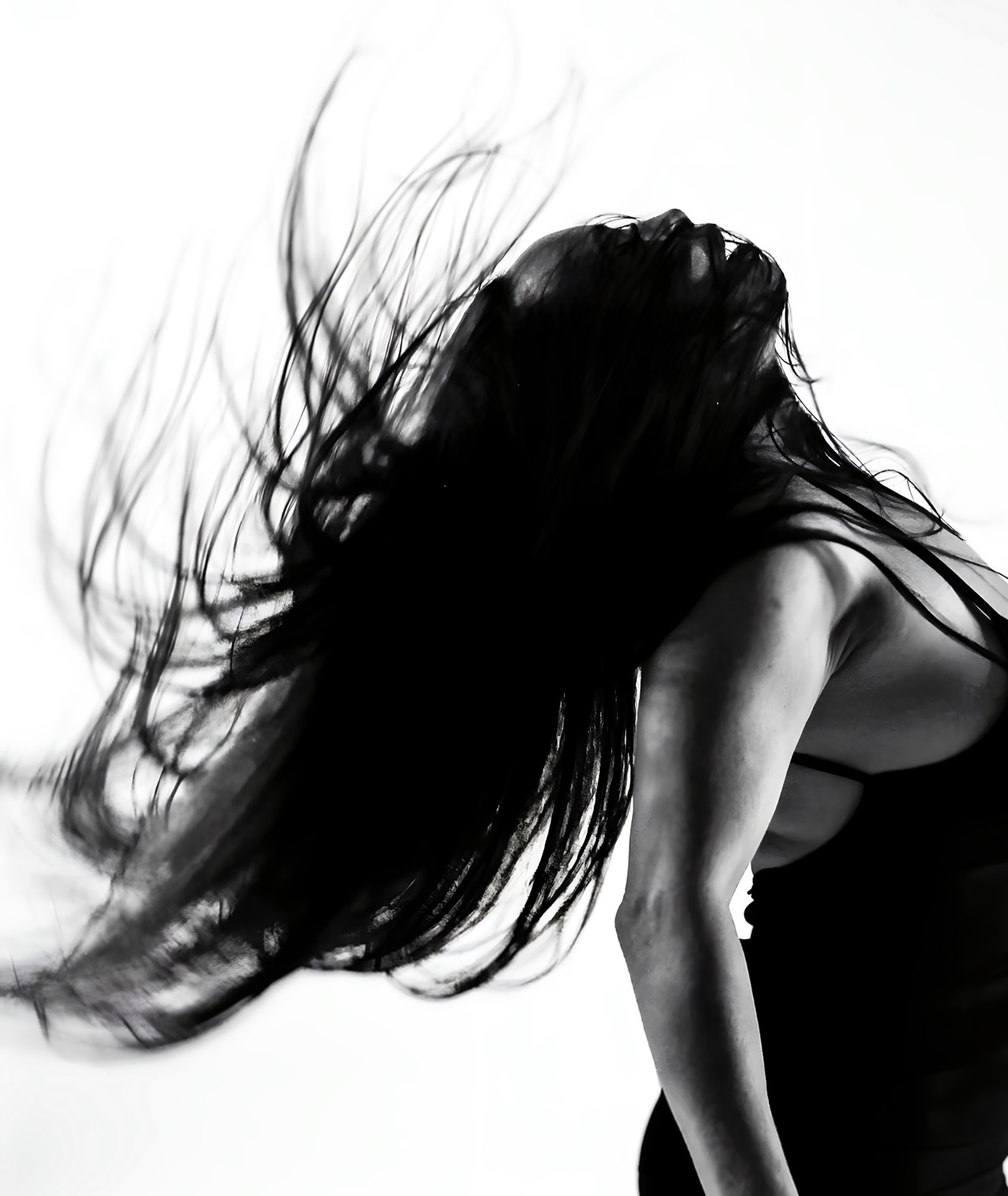 A high-contrast, black and white action shot captures Dr. Michelle Manu with her long, dark hair dramatically flowing upwards and backwards, creating a blurred effect. Her head is tilted back, looking towards the top right of the frame, and her face is mostly shadowed. She is wearing a dark, possibly strappy, top. The background is stark white.