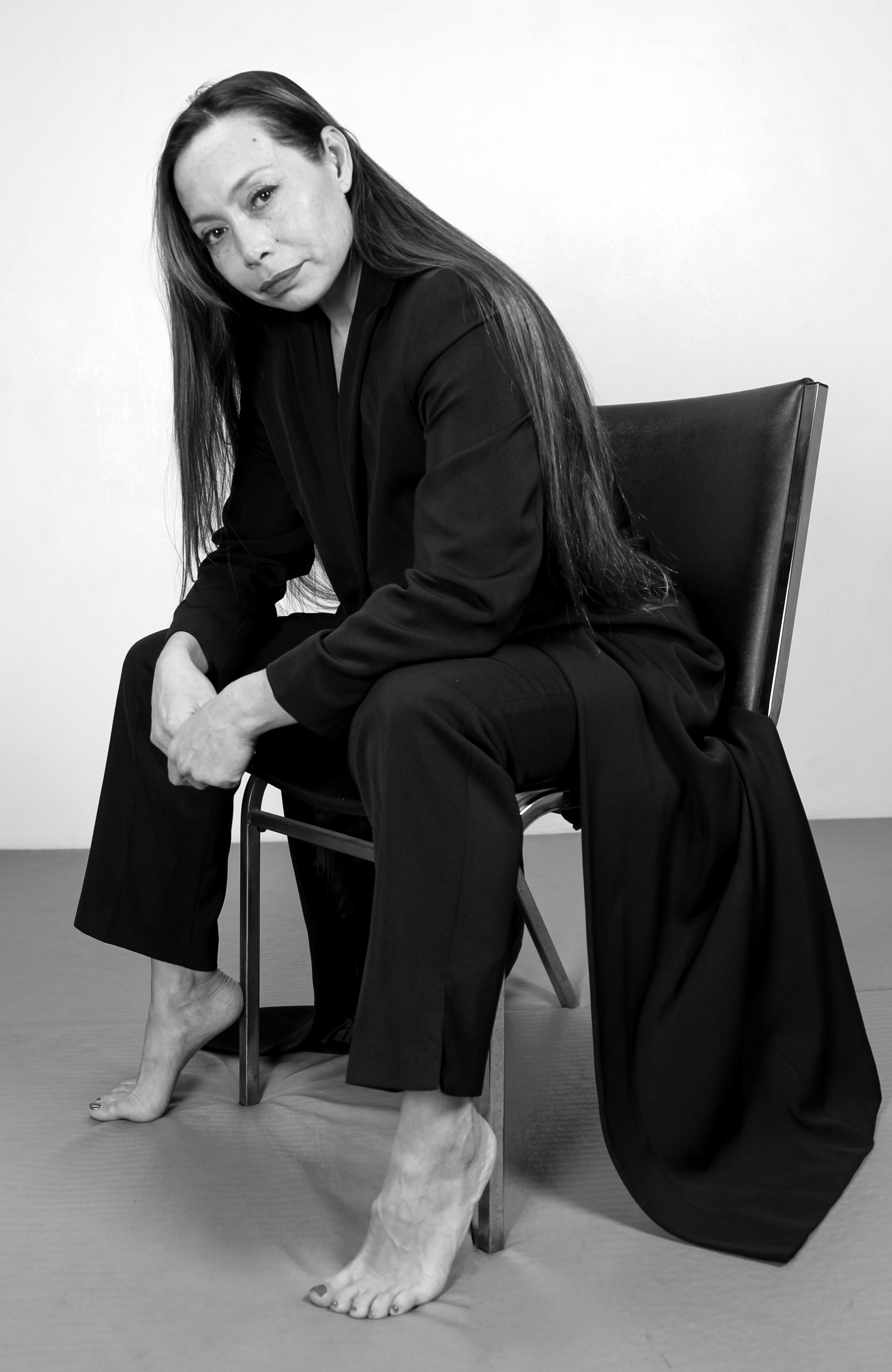 Black and white portrait of Michelle Manu, seated barefoot on a chair and dressed in a long, flowing black outfit. She leans slightly forward with her arms resting on her knees, gazing calmly at the camera. The minimalist background highlights her poised and introspective presence.