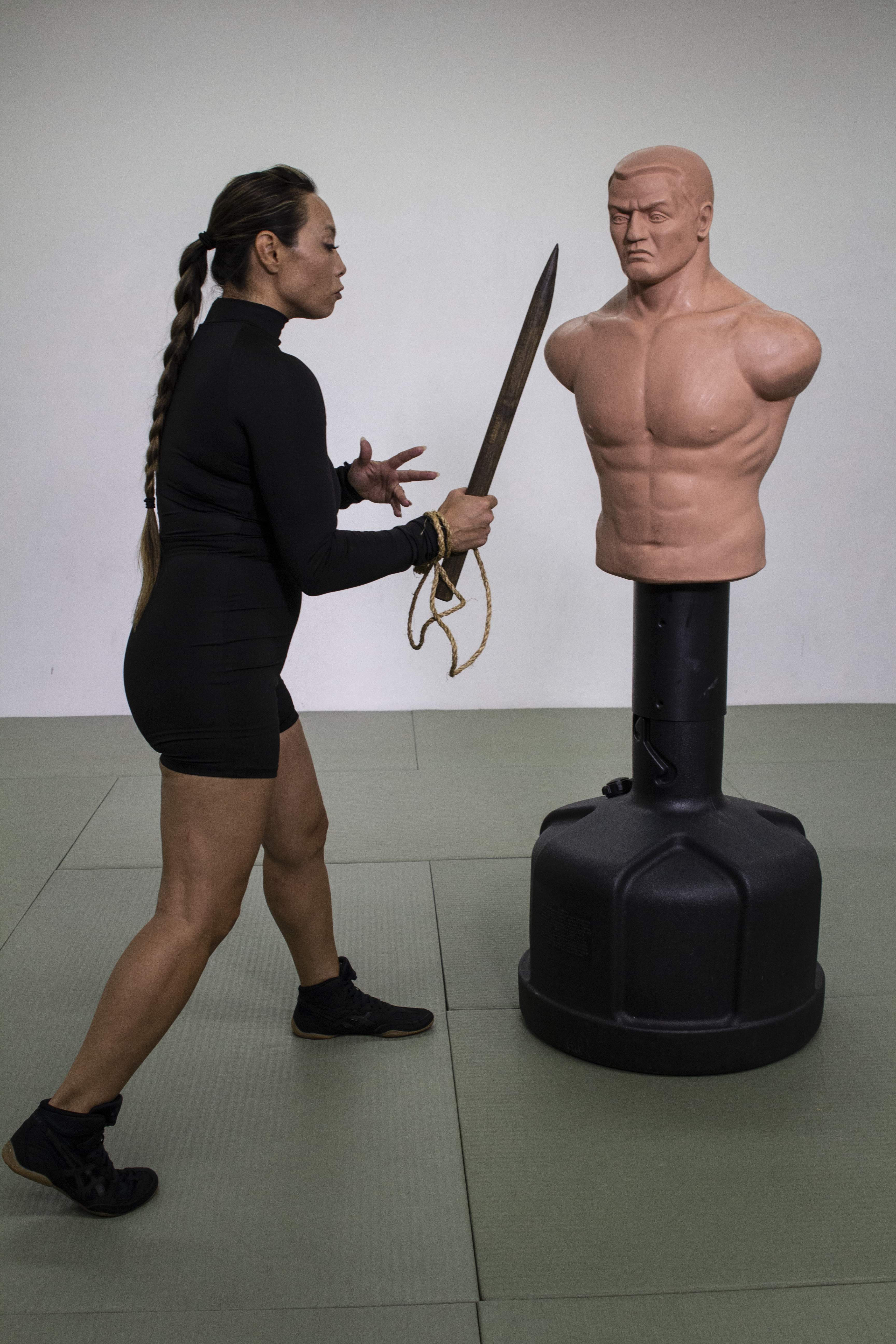 Kumu Michelle Manu, dressed in black athletic wear, practices a Pāhoa knife technique using a traditional-style training dummy with a muscular upper torso. She holds a wooden dagger in her right hand and a cord in her left, performing a controlled, ritualized movement on a dojo mat.