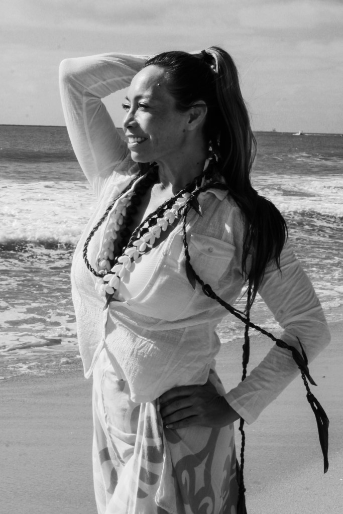 Dr. Michelle Manu stands on a beach with gentle waves in the background, looking off into the distance with a serene smile. She wears a light blouse, traditional Hawaiian lei, and a patterned wrap skirt. Her right hand rests on her hip while her left arm is raised to her head, with her long hair tied back in a ponytail. The black and white image captures a peaceful, reflective moment by the sea.