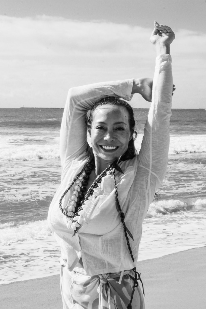 Dr. Michelle Manu smiles brightly while standing on a beach with waves rolling in behind her. She wears a light blouse, traditional Hawaiian lei, and patterned wrap skirt. Her arms are raised above her head in a joyful, relaxed stretch. The black and white photo captures a moment of serene celebration by the ocean.