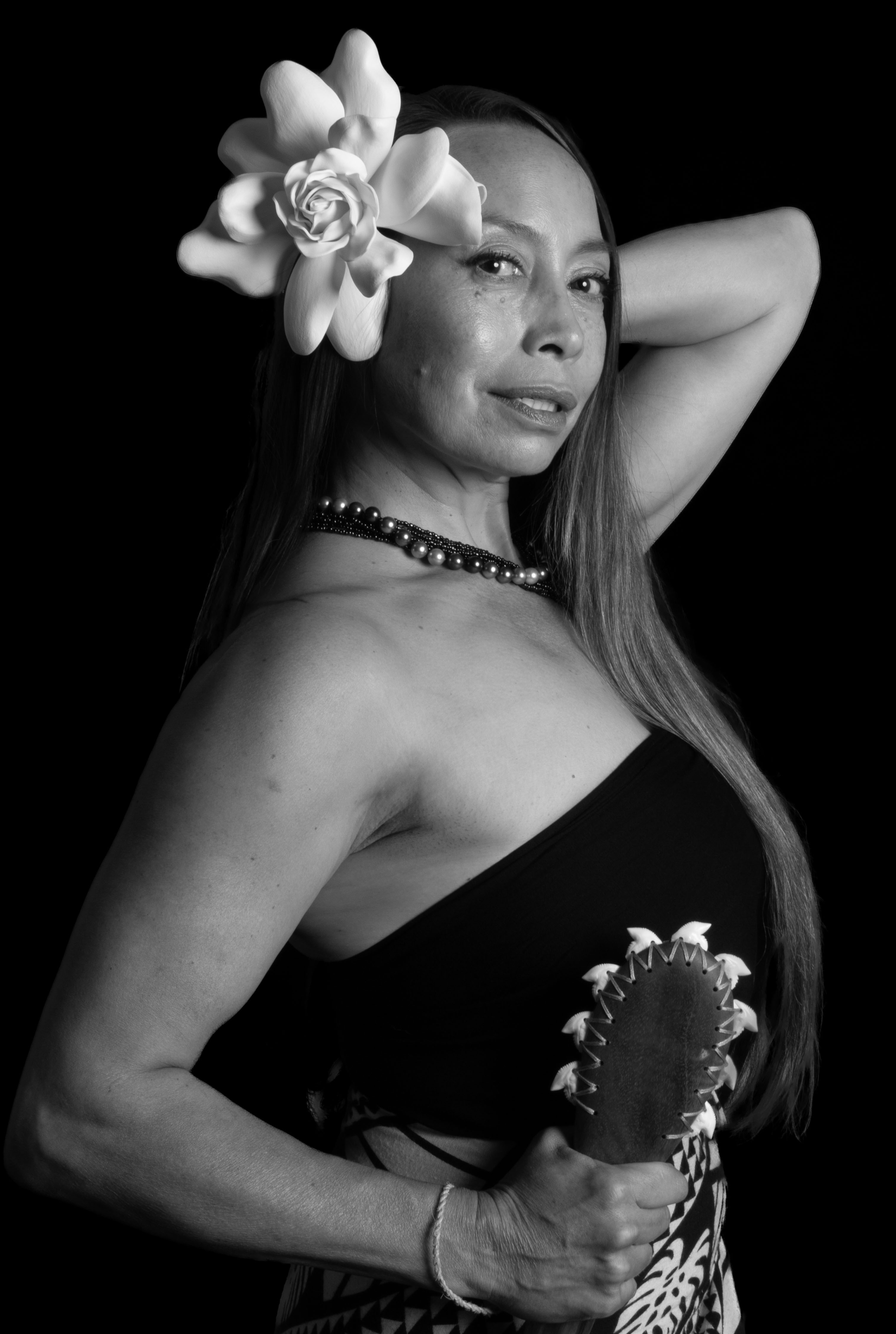 Dr. Michelle Manu poses gracefully in a black-and-white portrait, wearing a traditional garment and a large flower tucked behind her ear. She holds a Hawaiian kuʻekuʻe weapon—crafted with shark teeth—firmly in one hand. Her other arm is raised behind her head, and she wears a strand of black and white pearls around her neck. The image captures her strength, elegance, and cultural pride against a dark background.
