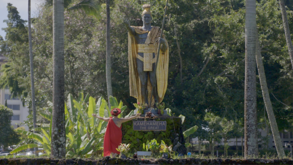 A long shot shows Dr. Michelle Manu, wearing a red haku lei (head lei) and a red dress, standing with her arms outstretched beside a large, golden statue of King Kamehameha the Great. The statue, depicting the king in a cloak and holding a spear, stands on a dark stone pedestal with "KAMEHAMEHA THE GREAT" inscribed on it. The scene is set outdoors with numerous tall palm trees and lush green foliage in the background, under a bright sky.