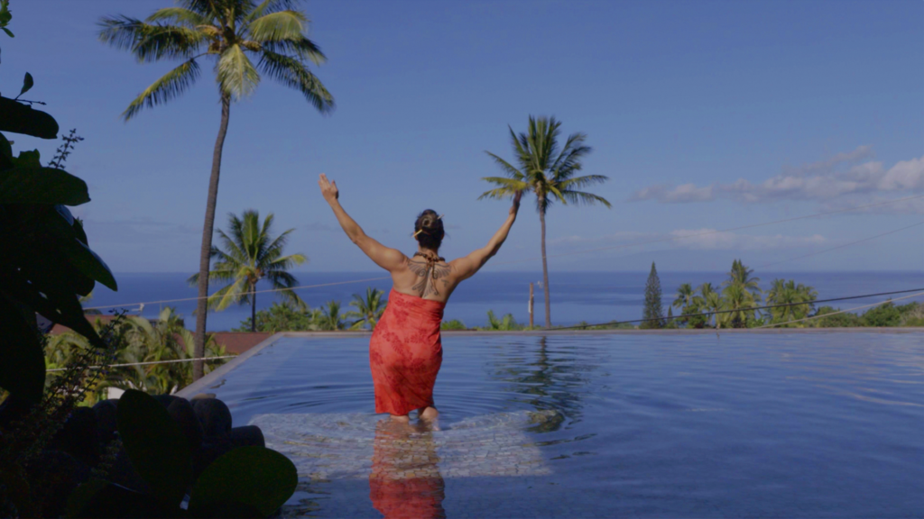 A full-back view of Dr. Michelle Manu shows her standing in an infinity pool, facing away from the camera, with her arms raised upwards. She is wearing a red sarong or dress and has a visible tattoo on her upper back. Beyond the pool, the vast blue Pacific Ocean stretches to the horizon under a clear sky. Several tall palm trees frame the scene, and lush green foliage is visible around the pool.