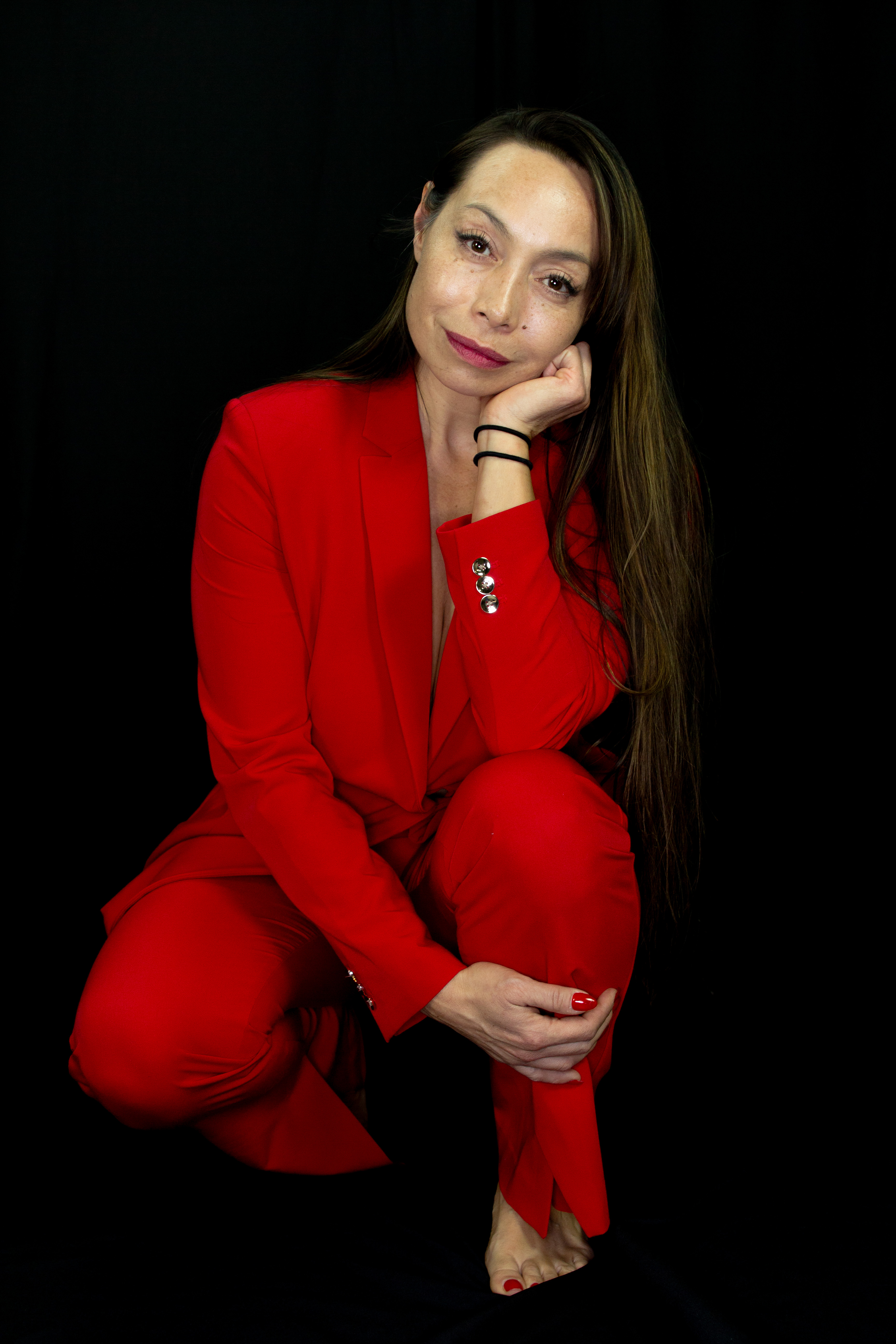 Michelle Manu poses barefoot against a black background, wearing a striking red suit. With long, straight hair, she crouches slightly, resting her chin thoughtfully on one hand while the other rests on her knee. Her nails are painted red to match her outfit, and her expression conveys calm confidence and introspection.