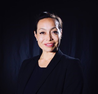 A headshot of Dr. Michelle Manu shows her smiling slightly, looking directly at the camera. She is a woman with light skin and dark hair pulled back. She is wearing a black blazer over a dark top. The background is a solid dark color, possibly black or dark grey.