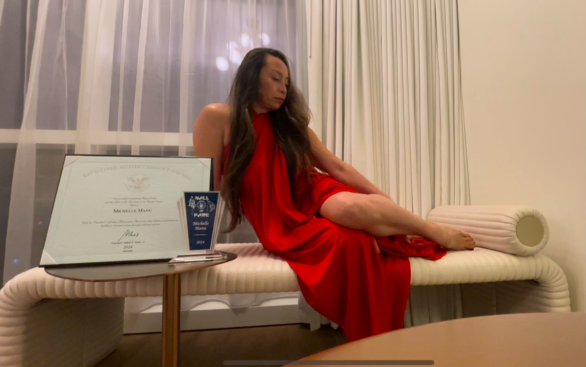 A woman identified as Dr. Michelle Manu is reclining on a light-colored, modern bench or chaise lounge. She is wearing a flowing red dress, and her long hair is draped over her shoulders. Her eyes appear to be closed or cast downwards. To her left, on a small round table, are a framed certificate, a small blue plaque or award, and what looks like a phone. The background features a window with sheer white curtains, suggesting an indoor setting