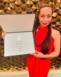 A medium shot of Dr. Michelle Manu shows her standing in front of a textured gold wall, smiling slightly and looking towards the viewer. She has long, dark hair pulled back from her face and is wearing a red, one-shoulder or strapless dress with a long, dark beaded necklace. She holds open a white certificate or diploma with both hands, positioned to be clearly visible. Her right wrist sports a gold watch.
