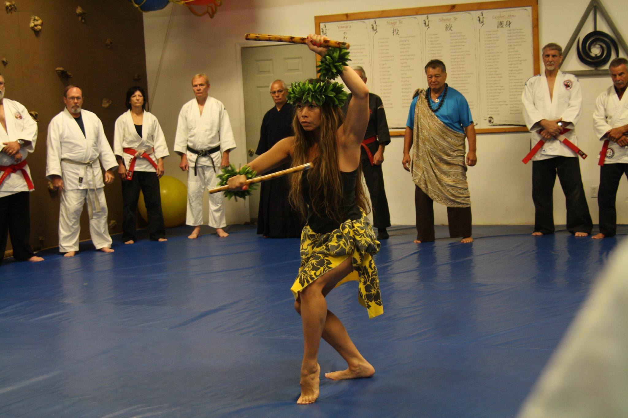 Dr. Michelle Manu performs a traditional Hawaiian martial arts demonstration using wooden sticks, adorned with leafy adornments on her head and wrists. She wears a yellow and black patterned skirt and a black top. A group of martial artists in uniforms and traditional attire observe respectfully in the background, standing on a blue mat inside a dojo.