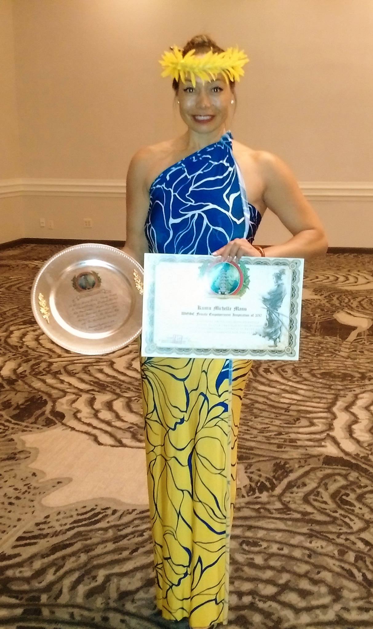 Dr. Michelle Manu stands smiling in a brightly patterned traditional Hawaiian outfit, wearing a yellow flower crown. She holds a certificate and a silver award plate, indicating recognition or an honor received. The setting appears to be a formal indoor venue with patterned carpet and neutral walls.
