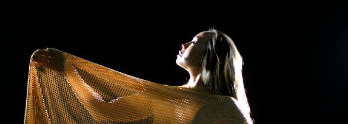 Dr. Michelle Manu is illuminated dramatically against a dark background, facing upward with closed eyes in a serene pose. She is draped in a golden, mesh-like fabric that flows over her arms, evoking a sense of elegance, power, and spiritual expression.