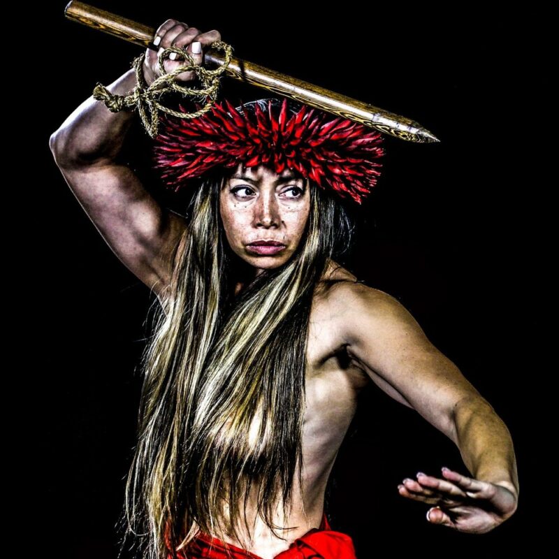 Dr. Michelle Manu wearing a traditional red feather headdress and garment, holding a wooden spear overhead with one arm extended in a poised martial stance against a black background.