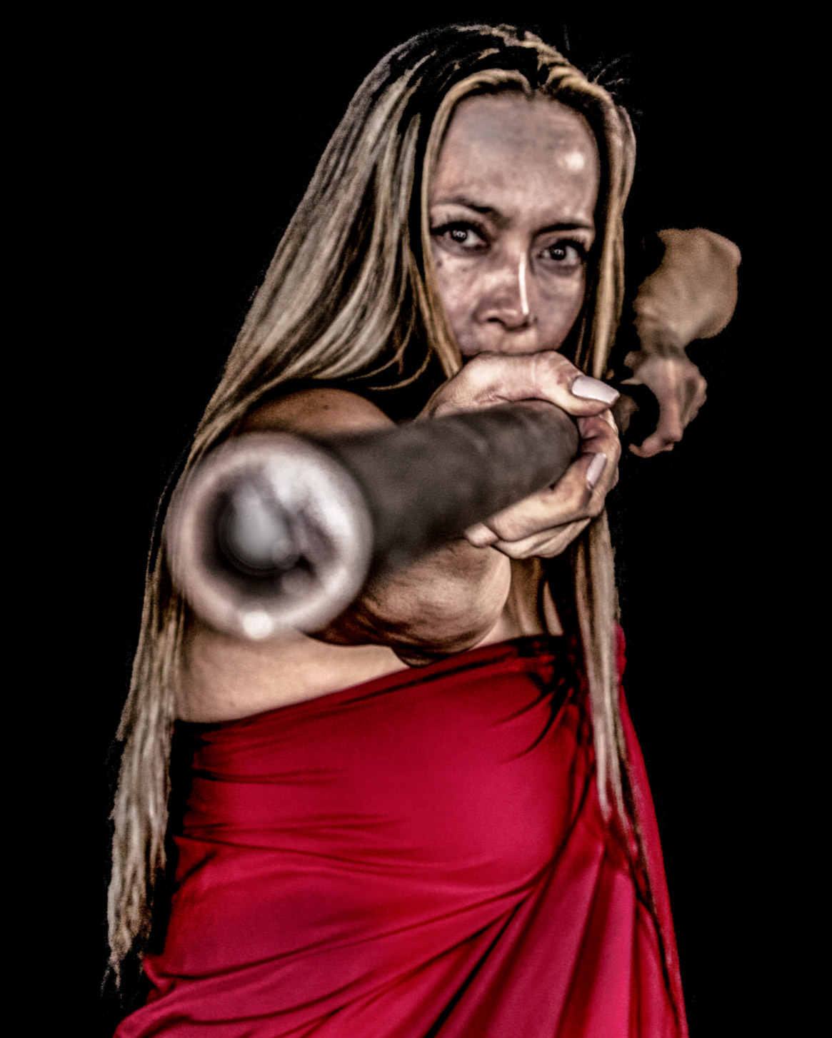 Dr. Michelle Manu, wrapped in a red garment, holds a long staff aimed directly at the camera with an intense, focused expression against a dark background.