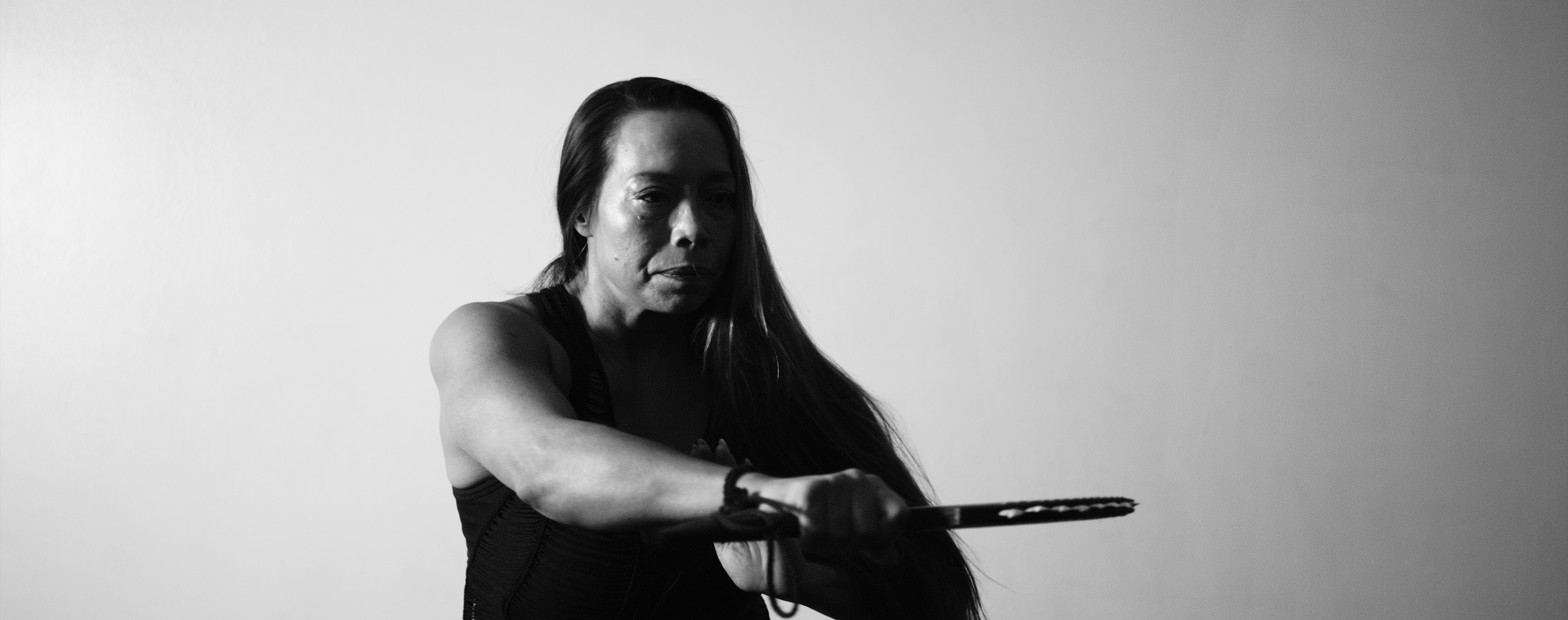 Black-and-white photo of Dr. Michelle Manu, wearing a sleeveless top, focused and holding a long-bladed sword in both hands during a martial arts stance.