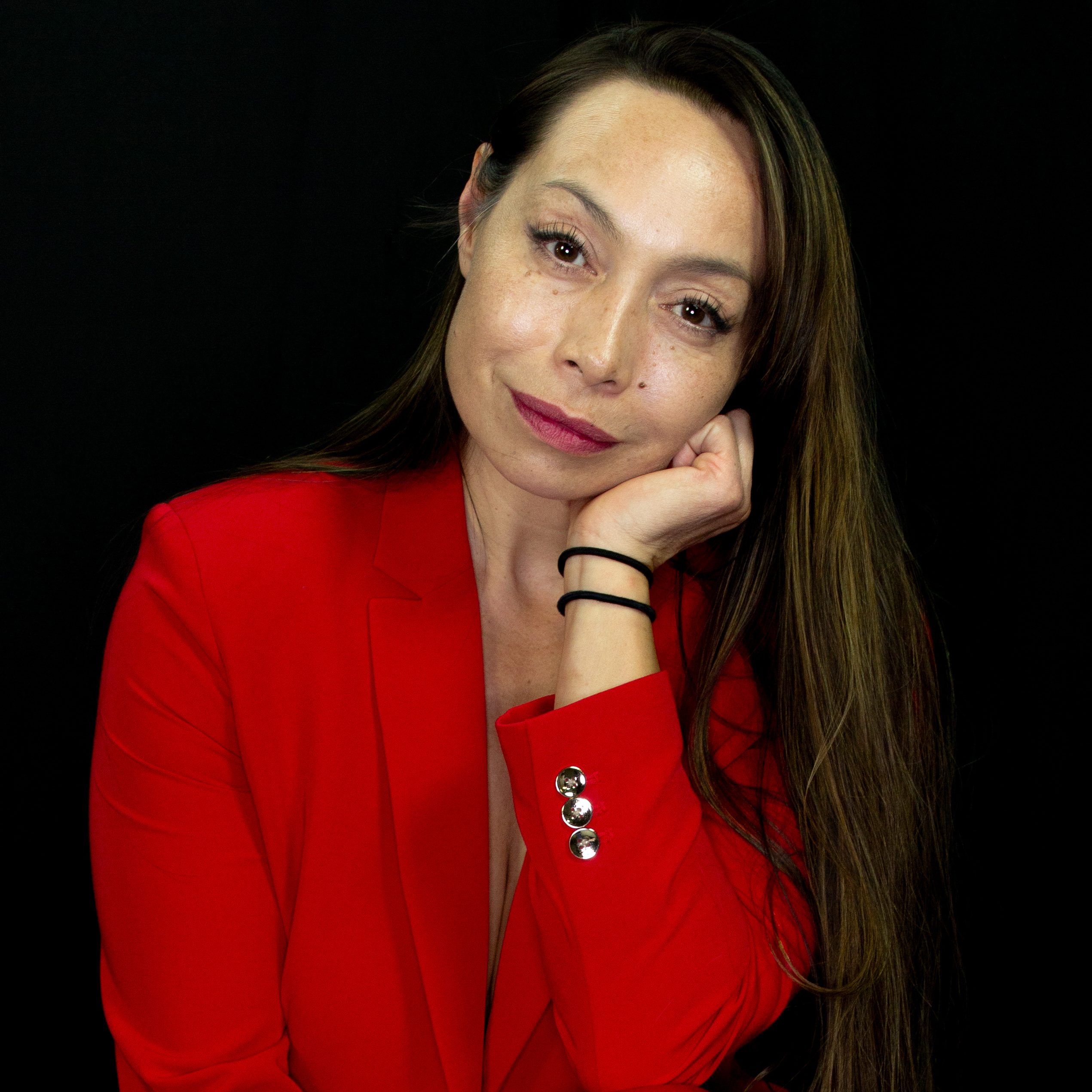 Michelle Manu poses barefoot against a black background, wearing a striking red suit. With long, straight hair, she crouches slightly, resting her chin thoughtfully on one hand while the other rests on her knee. Her nails are painted red to match her outfit, and her expression conveys calm confidence and introspection.